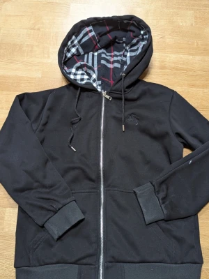 Svart hoodie med rutigt foder - Säljer en svart hoodie med dragkedja från Burberry. Hoodien har ett rutigt foder i huvan med grå, vit och röd design, samt ribbade muddar och fickor framtill. Materialet är mjukt och bekvämt, perfekt för en avslappnad stil.