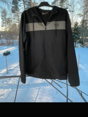 Svart vindjacka från Under Armour - Snygg svart vindjacka från Under Armour med dragkedja framtill och huva. Jackan har en grå panel över bröstet med logga på vänster sida. Perfekt för blåsiga dagar och enkel att matcha med olika outfits.