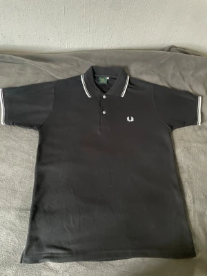 Svart piké från Fred Perry - Klassisk svart pikétröja från Fred Perry med vit logga på bröstet och vita ränder på krage och ärmslut. Tröjan har korta ärmar, knappar vid halsen och är gjord i bomull för en skön känsla. Perfekt till en clean och stilren look. Använd 2-4 gånger och är i mycket fint skick.