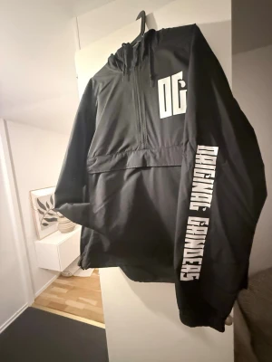 Original grinders vindjacka med tryck - Svart vindjacka från original grinders med stor vit logga på bröstet och texten 'original grinders ' i vitt längs ena ärmen. Jackan har huva med dragsko och halv dragkedja framtill. Tillverkad i 100% polyester, perfekt för streetwear-stil.