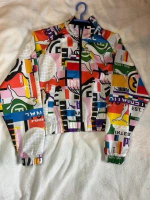 Färgglad vindjacka från Puma 2021 - OANVÄND! Säljer en supercool vindjacka från Puma år 2021 med färgstarkt, grafiskt mönster och loggor i flera färger. Jackan har dragkedja framtill, hög krage och resår vid ärmslut och midja. Perfekt för dig som vill sticka ut med din stil!