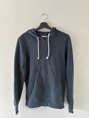 Mörkblå hoodie med vita snören - En stilren mörkblå hoodie med vita dragsnören och stor känguruficka framtill. Hoodien har en klassisk passform och är tillverkad i mjukt bomullsmaterial. Perfekt för en avslappnad look och enkel att matcha med jeans eller joggers.