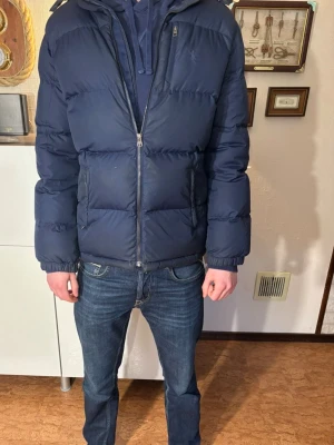 Mörkblå pufferjacka från Ralph lauren  - Säljer en mörkblå pufferjacka med stor huva och quiltad design. Jackan har dragkedja framtill och sidofickor, perfekt för kalla vinterdagar. Materialet är syntet och jackan är tjock och isolerande. Passar dig som vill ha en stilren och varm vinterjacka. Märke Ralph lauren 