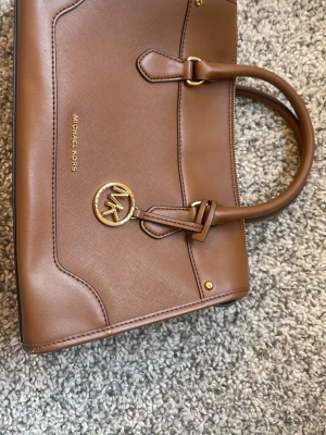 Brun handväska från Michael Kors - Snygg brun handväska från Michael Kors i slätt läder med guldfärgade detaljer och logotyp. Väskan har dubbla handtag och en klassisk, stilren design med dekorativ MK-berlock. Perfekt accessoar för dig som gillar tidlös och elegant stil. Inga slitningar eller liknande!