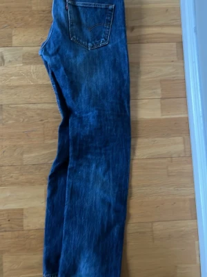 Levi's 501 raka blå jeans - Klassiska Levi's 501 jeans i mörkblå denim med rak passform. Jeansen har fem fickor, knappgylf och den ikoniska läderpatchen bak i midjan. Perfekta för en avslappnad och tidlös stil.