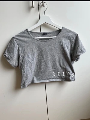 Grå croppad t-shirt med tryck - Grå croppad t-shirt från Reload med rund halsringning och korta ärmar. T-shirten har ett vitt tryck med texten 'RELOAD' nertill. Perfekt för en avslappnad och trendig stil.