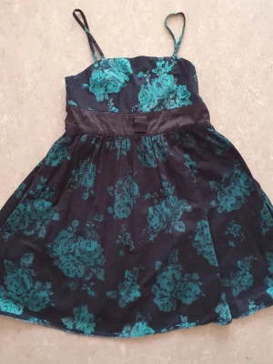 Klänning - Snygg svart klänning med turkos blommönster och smala axelband. Klänningen har en markerad midja med ett brett band och lätt utställd kjol. Perfekt för dig som gillar en feminin och trendig stil. Finns fläck.