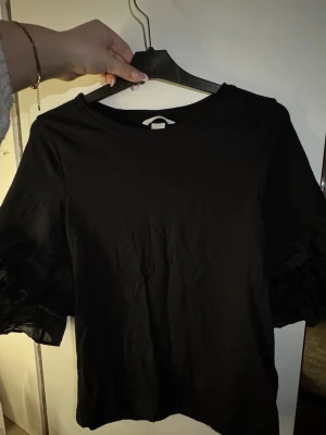 Svart topp med volangärmar - Snygg svart topp med rund hals och unika volangdetaljer på ärmarna. Tillverkad i mjukt material med transparenta inslag på ärmarna som ger en cool touch. Perfekt för dig som vill ha något extra till outfiten.