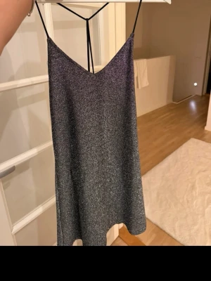 Glittrig silver kortklänning - Snygg glittrig silverfärgad kortklänning med tunna axelband och v-ringad hals. Klänningen har en enkel, rak passform och är ärmlös, perfekt för att sticka ut på festen. Materialet känns mjukt och skimrande.