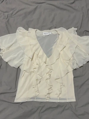 Krämvit volangblus från Kappahl - Söt krämvit blus från Kappahl med v-ringning och stora volanger framtill och på ärmarna. Tillverkad i ett tunt, transparent material som ger en luftig känsla. Perfekt för dig som gillar romantisk och trendig stil.