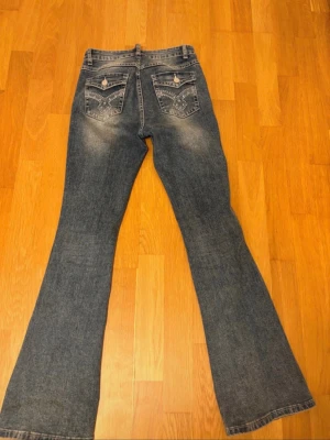 Blå bootcut jeans med detaljer - Säljer ett par blå bootcut jeans med snygga detaljer på bakfickorna. Jeansen har mid waist och klassisk femficksmodell. Passformen är utsvängd nedtill för en retro vibe.