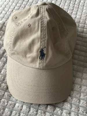 Beige keps från Polo Ralph Lauren - Snygg beige keps från Polo Ralph Lauren med klassisk böjd skärm och mörkblå broderad logga framtill. Tillverkad i bomullstyg och har ventilerande hål upptill för extra komfort. Perfekt accessoar för en avslappnad och stilren look.