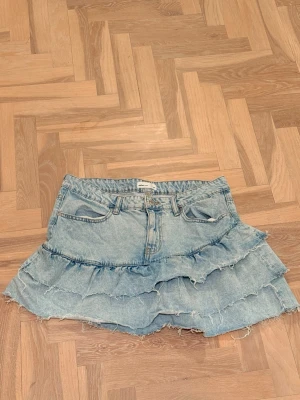 Ljusblå jeanskjol med volanger - Trendig ljusblå jeanskjol med två lager volanger och råa kanter. Kjolen har klassiska fickor fram och bak samt bälteshällor. Från Gina Tricot