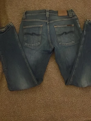 Blå jeans från Nudie Jeans - Säljer ett par klassiska blå jeans från Nudie Jeans med raka ben och snygga slitningar. Jeansen har fem fickor, ljus tvätt och den ikoniska vågdetaljen på bakfickorna. Perfekta för en avslappnad och trendig stil. Hör av er vid minsta fråga!