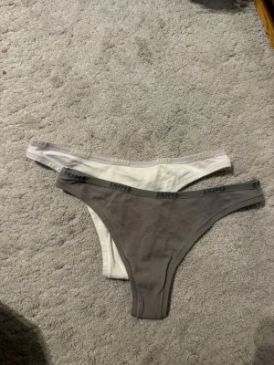 2-pack stringtrosor från Sloggi - Två par stringtrosor från skims i mjuk bomullsblandning. Ena paret är grått och det andra vitt, båda med resårkant där Sloggi-loggan syns runtom. Klassisk stringmodell som sitter skönt och diskret under kläder.