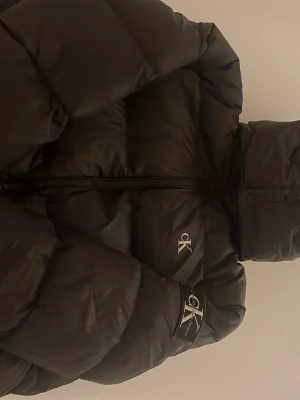 Svart pufferjacka från Calvin Klein storlek M - Storlek:M. Säljer en svart pufferjacka från Calvin Klein med huva och dragkedja framtill. Jackan har logga på bröstet och ärmen, samt breda, vadderade paneler för extra värme. Perfekt för kalla vinterdagar och riktigt snygg streetstil.