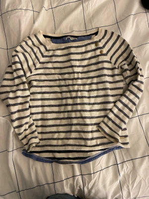 Randig långärmad tröja med knappar - Snygg långärmad tröja med blå och vita horisontella ränder. Tröjan har båtringning och tre dekorativa knappar vid axeln. 