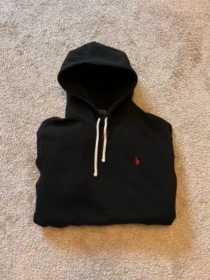 Svart hoodie från Polo Ralph Lauren - Svart hoodie från Polo Ralph Lauren med vit dragsko och klassisk röd logga broderad på bröstet. Mjuk bomullskänsla och huva gör den perfekt för en chill och stilren look.