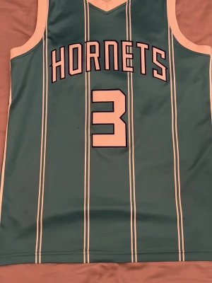 Hornets basketlinne Rozier III #3 - Snyggt turkost basketlinne från Hornets med vita vertikala ränder och vita detaljer. Framsidan har texten 'HORNETS' och siffran 3, baksidan har 'ROZIER III' och samma nummer. Perfekt för basketfans som vill sticka ut på planen. Storlek M