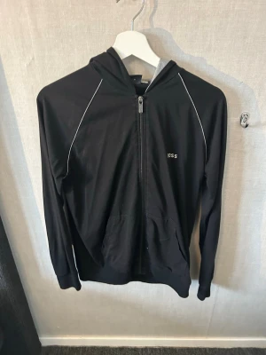 Svart zip hoodie från BOSS - Snygg svart hoodie från BOSS med dragkedja framtill och vit piping längs axlarna. Huva med grå insida och fickor på magen. Minimalistisk logga på bröstet. Perfekt för en clean och sportig look.