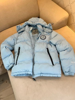 Ljusblå pufferjacka från Lorenzo - Snygg ljusblå pufferjacka från Lorenzo med huva och två svarta dragkedjefickor framtill. Jackan har Lorenzo-logga på bröstet och justerbar dragsko i huvan. Perfekt för kalla vinterdagar och riktigt trendig look. Lorenzo är ett märke från UK och inte så kännt här i sverige så hör av er om ni har frågor osv! 