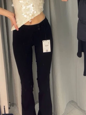 Svarta bootcut kostymbyxor  med låg midja - Snygga svarta kostymbyxor med bootcut-modell och låg midja. Byxorna har knäppning framtill och sitter tajt över höfterna innan de blir vidare nedtill. Perfekta för dig som gillar en klassisk men trendig look.