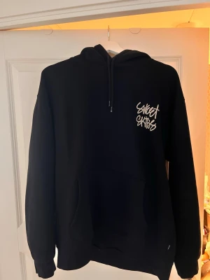 Svart hoodie Sweet SkTBS - Svart hoodie från Sweet SkTBS med stor vit logga i graffiti-stil på ryggen och mindre logga på bröstet. Klassisk känguruficka framtill och justerbar huva med snören. Mjuk bomullsblandning som känns skön mot huden. Perfekt för en avslappnad streetwear-look.