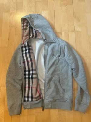 Grå burberry hoodie  - Snygg grå hoodie från Burberry med dragkedja och klassiskt rutigt foder i huvan. Mjukt material och två fickor framtill. Perfekt för dig som gillar stilrena plagg med en twist av lyx. Huvan har dragsko och insidan är fodrad i vitt teddytyg.