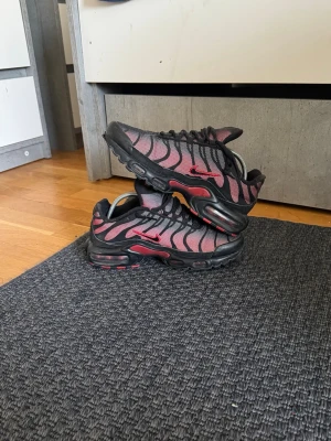 Nike Air Max Plus TN svart/röd - Nike Air Max Plus TN sneakers med svart och röd mesh och syntetmaterial. Skorna har en ikonisk vågig design, synlig Air Max-dämpning i sulan och röda detaljer som Swoosh-loggan. Perfekt för dig som gillar streetstyle och sportiga vibes.