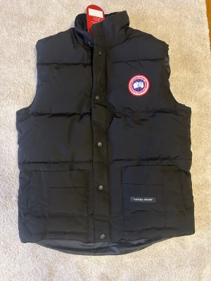 Svart dunväst från Canada Goose - Stilren svart dunväst från Canada Goose med klassisk logotyp på bröstet. Västen har hög krage, tryckknappar framtill och två stora fickor med Canada Goose-märke. Perfekt för kalla dagar och snygg att kombinera med hoodie eller tröja.