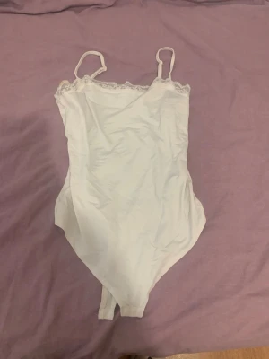 Vit body med spetsdetaljer - Säljer en vit body med tunna axelband och fina spetsdetaljer längs halsringningen. Bodyn har en enkel och stilren design, perfekt för att matcha med olika outfits. Materialet känns mjukt och bekvämt mot huden.