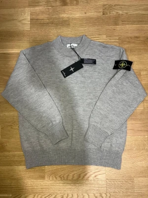 Stone Island Tröja - Stickad tröja från Stone Island i klassisk ljusgrå färg. Rund hals, ribbade muddar och nederkant samt den ikoniska Stone Island-badgen på ärmen. Storlek M med normal passform. Helt ny, aldrig använd. Originaltags medföljer. Skickas snabbt och säkert vid affär.