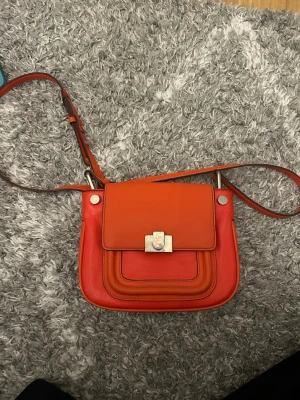 Orange axelväska i läder - Snygg axelväska i orange läder med justerbar axelrem och silverfärgat spänne framtill. Väskan har ett lock och flera lager på framsidan som ger en cool look. Perfekt för dig som vill ha en färgklick till outfiten.