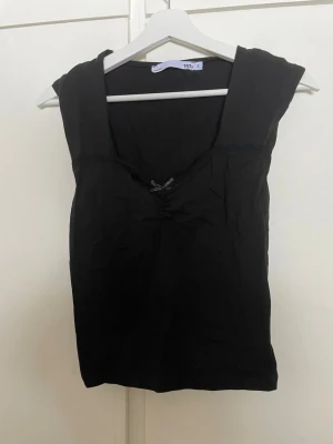 Svart ärmlös topp från 157 - Snygg svart ärmlös topp från 157 med v-ringning och liten rosett framtill. Toppen har en enkel och stilren design som passar till många olika outfits. Perfekt för dig som gillar minimalistisk stil och vill ha något bekvämt och lättmatchat.