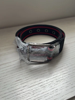 Svart och rött bälte med metallspänne - Snyggt svart bälte med röda detaljer och silverfärgat metallspänne. Bältet har flera metallöljetter längs hela längden och är gjort i slitstarkt syntetmaterial. Perfekt för att ge din outfit en cool touch.