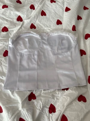 Vit axelbandslös korsett med spets - Snygg vit axelbandslös korsett med bygel och spetsdetaljer upptill. Toppen har en silkeslen finish och dragkedja bak för enkel påklädning. Perfekt för att styla med höga jeans eller kjol för en trendig look. Defekter finns! Den är nopprig som syns på de två sista bilderna men det är inget man tänker på när den är på! Även så har någon enstaka söm gått av men det är inte heller något som syns på!💓🌸