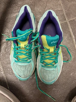 Turkosa Asics sneakers med gula detaljer - Snygga sneakers från Asics i turkos mesh med gula och gröna detaljer. Skorna har gröna snören, vit och grön sula samt lila insida. Perfekta för dig som vill ha färgglada och bekväma sneakers till vardags. Klassisk Asics-logga på sidan. Slitna längst fram på ena skon. 