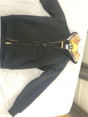 Svart hoodie med Burberry-detaljer - Snygg svart hoodie med dragkedja från Burberry. Klassisk huva med det ikoniska Burberry-rutiga mönstret på insidan och justerbara snören. Två stora fickor framtill och guldfärgade dragkedjedetaljer. Perfekt för en trendig och avslappnad stil.