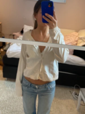 Vit croppad kofta med knappar - Säljer en vit croppad kofta med knappar framtill och långa ärmar. Koftan har en enkel design och är perfekt att slänga på över ett linne eller t-shirt. Materialet ser ut att vara mjukt och lätt, vilket gör den bekväm att bära.