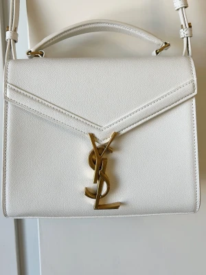 Cassandra mini trendig väska - Cassandra mini i vitt/cremefärgat kalvskinn. Så kallat "grained" läder vilket gör att eventuella märken bara är att torka bort. Mått ca längd 20 höjd 16 djup 7.5cm. Top handle men axelband medföljer så att man kan bära den på flera sätt. Använd en gång så i nyskick. 