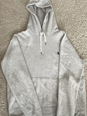 Ralph Lauren hoodie - Snygg ljusgrå hoodie från Polo Ralph Lauren med klassisk huva och vita dragsnören. Har en känguruficka framtill och den ikoniska mörkblå loggan broderad på bröstet. Tillverkad i mjuk bomullsblandning som känns skön mot huden. Perfekt för en avslappnad och stilren look.