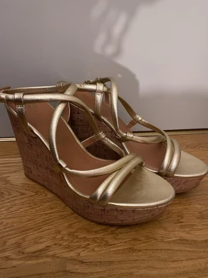 Guldiga kilklackade sandaler - Snygga sandaler med kilklack i kork och metallic guldiga remmar. Öppen tå och korsade band över foten ger en trendig look. Perfekta för sommarens alla tillfällen när du vill sticka ut lite extra.