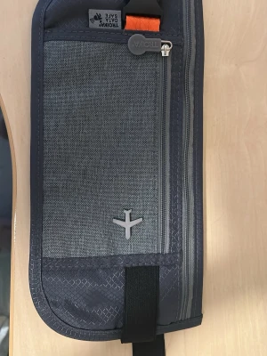 Grå Troika plånbok med dragkedja - Snygg och praktisk grå plånbok från Troika med RFID-skydd. Plånboken har flera fack, dragkedja och en svart spänne med orange detalj. Tillverkad i slitstarkt tygmaterial och har en stilren design med ett flygplansmotiv på framsidan.