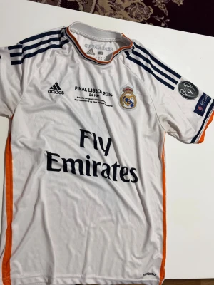 Real Madrid Ronaldo tröja 2014 - Vit Real Madrid matchtröja från Champions League-finalen 2014 med Ronaldo 7 på ryggen. Tröjan har svarta detaljer, orange sidor, klubbmärke, Adidas-logga och Fly Emirates-tryck. Materialet är lätt och andas, perfekt för fotbollsträning eller match.