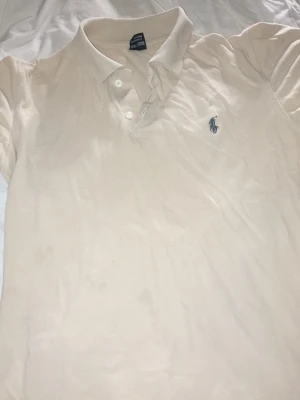 Beige pikétröja från Ralph Lauren - Klassisk beige pikétröja från Ralph Lauren med den ikoniska broderade loggan på bröstet. Tröjan har krage, knappar framtill och är tillverkad i bomull för en skön och avslappnad stil. Strlk M sitter lite som S,M. Litet litet hål som du ser på sista bilden