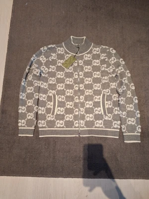 Grå zip-kofta med GG-mönster från Gucci - Snygg grå kofta med vit GG-logotyp över hela plagget från Gucci. Koftan har dragkedja framtill, ribbade muddar och krage samt två fickor. Materialet är stickat och känns lyxigt och mjukt. Perfekt för dig som gillar exklusiva märken och stilrena detaljer.