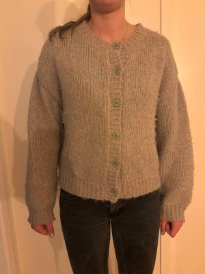 Ljus grön och rosa stickad kofta med knappar - Mysig grön och rosa stickad kofta med stora gröna knappar framtill. Koftan har en rak passform, ribbade muddar vid ärmslut och nederkant samt rund halsringning. Perfekt för lager på lager och enkel att matcha med olika outfits.