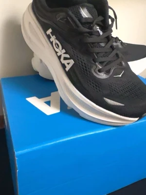 Svarta Hoka sneakers med vit sula - Snygga svarta Hoka sneakers med tjock vit sula och ventilerande mesh-ovandel. Skorna har klassisk låg profil, vita detaljer och Hoka-logga på sidan. Perfekta för dig som gillar sportig och modern stil.