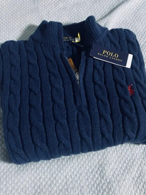 Mörkblå kabelstickad tröja Polo Ralph Lauren - Mörkblå kabelstickad tröja från Polo Ralph Lauren med halv dragkedja och klassisk röd logga broderad på bröstet. Tröjan har hög krage och är tillverkad i mjukt material som ger en lyxig känsla. Perfekt för dig som gillar stilrena och tidlösa plagg.