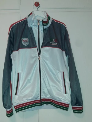 Everlast retro träningsjacka Italy edition - Snygg retro träningsjacka från Everlast i vitt, grått och rött med gröna detaljer. Jackan har dragkedja, ståkrage och ribbade muddar med ränder. Två fickor fram och broderad logga på bröstet. Perfekt för sport eller streetwear. Stolek  för man, storlek M för kvinna.